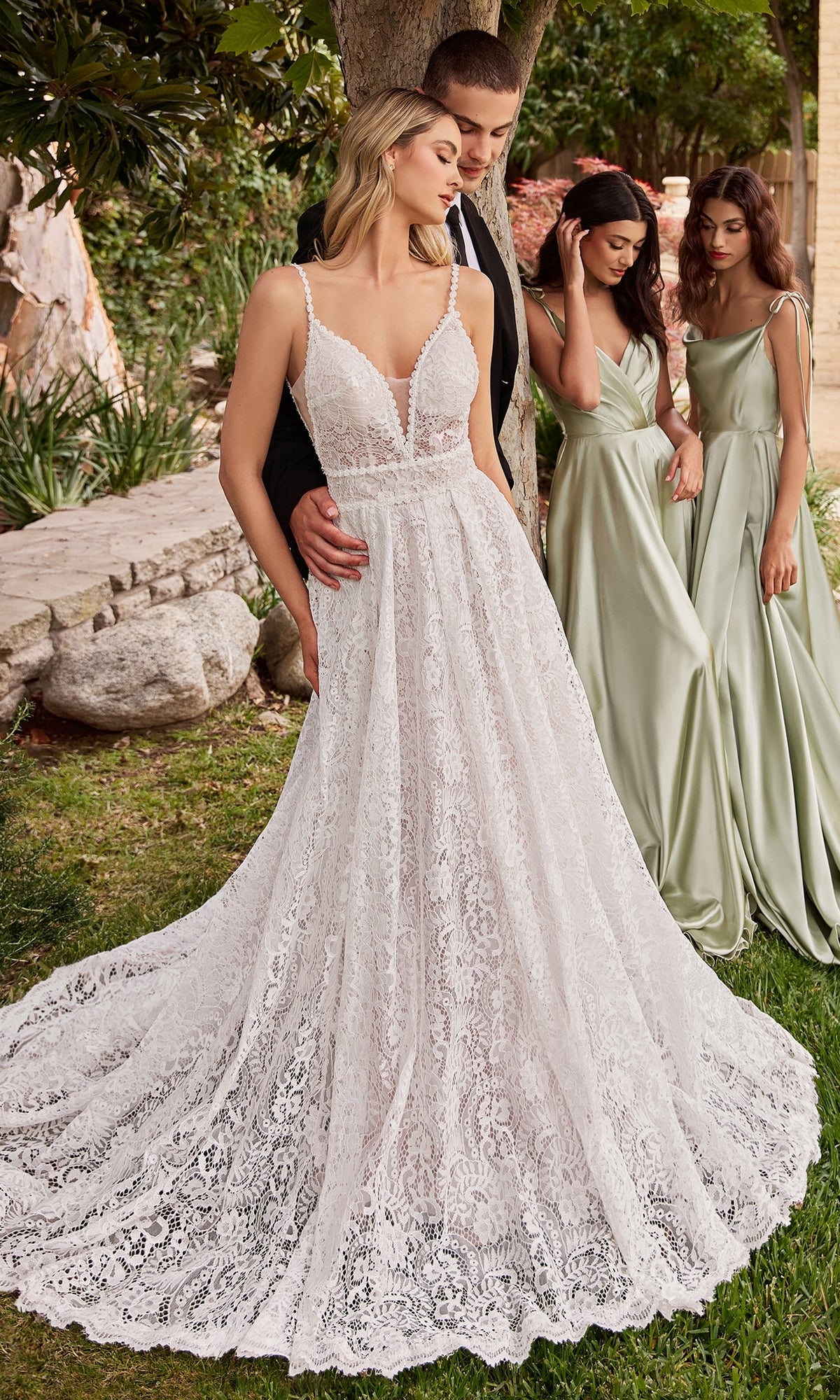A bride in the Ladivine A-Line Lace Wedding Dress CD862W stands with the groom, while two bridesmaids in sage green dresses stand nearby outdoors.